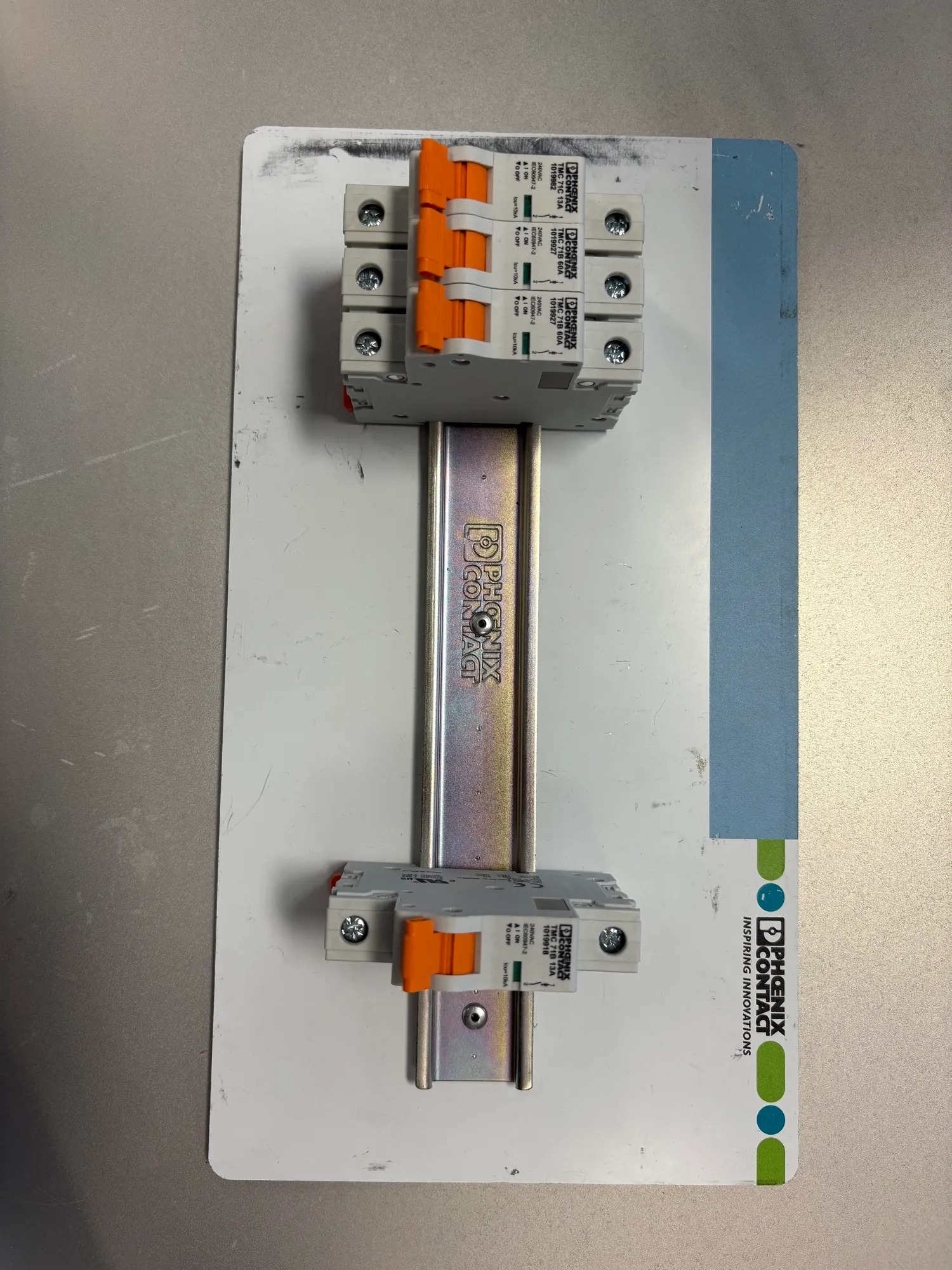 Phoenix Contact circuit breakers with orange switches mounted on a DIN rail, with three grouped at one end and one at the other, on a white base plate.