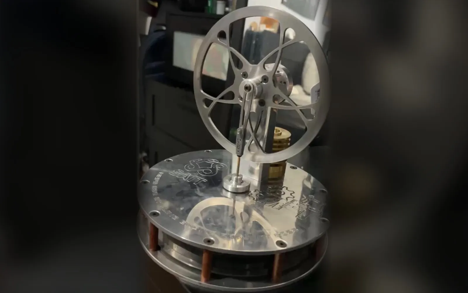 Finished Stirling engine with a machined aluminum flywheel and connecting rod on a round aluminum plate engraved with the SAIT logo, standing on copper posts.